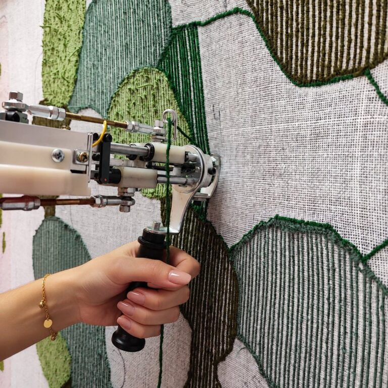 Mão segurando a tufting gun e aplicando fio verde num tecido com desenho de folhas